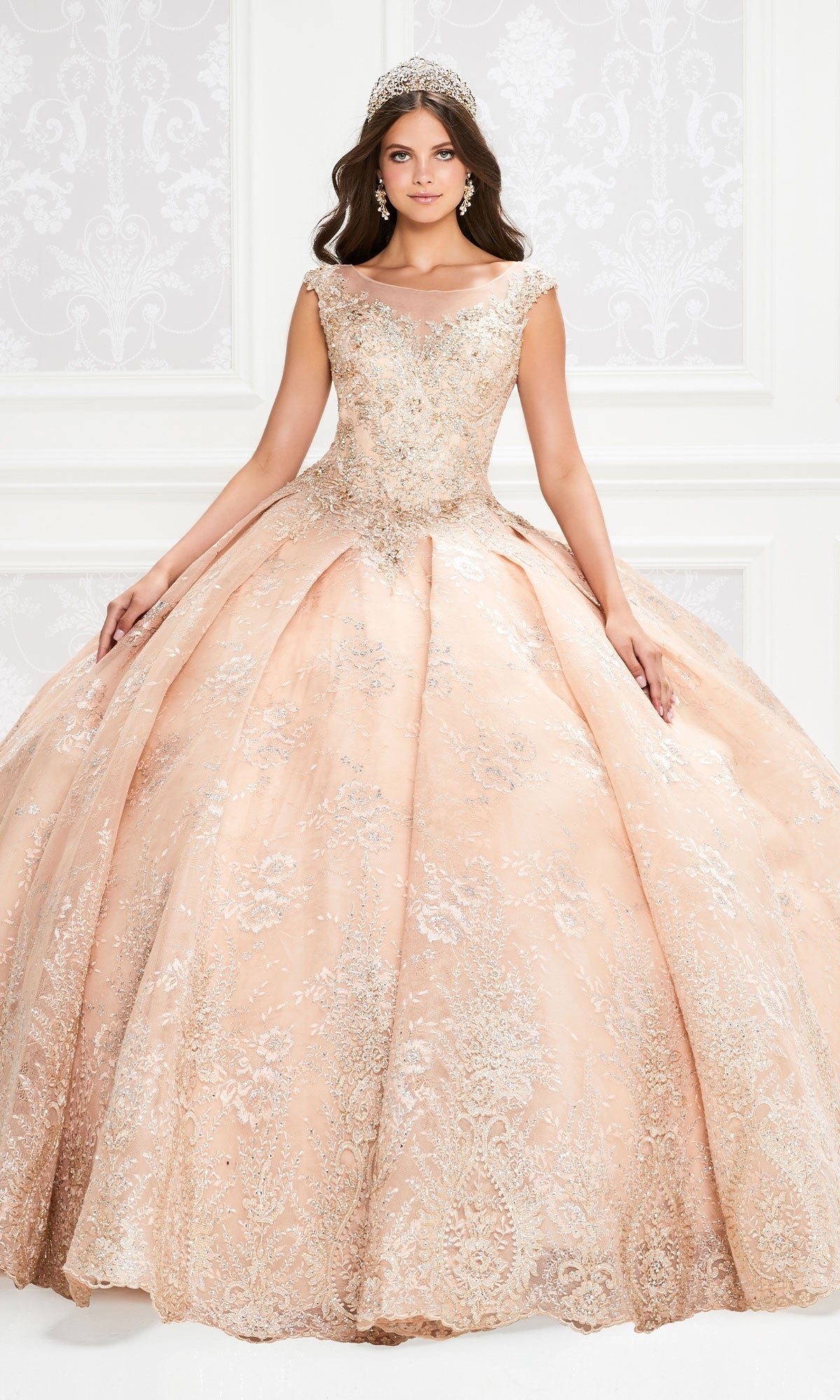 A woman in the Princesa Traditional Lace Quinceañera Dress PR12002, a peach ball gown with silver embellishments and a tiara, stands before an ornate white backdrop.